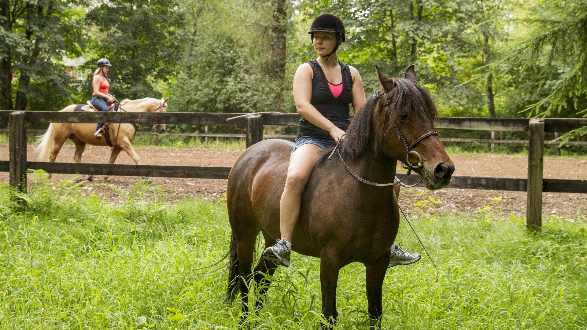 A women sits bare back on a horse in a patch of overgrown grass. There is a fence behind them and another rider on the other side of the fence.